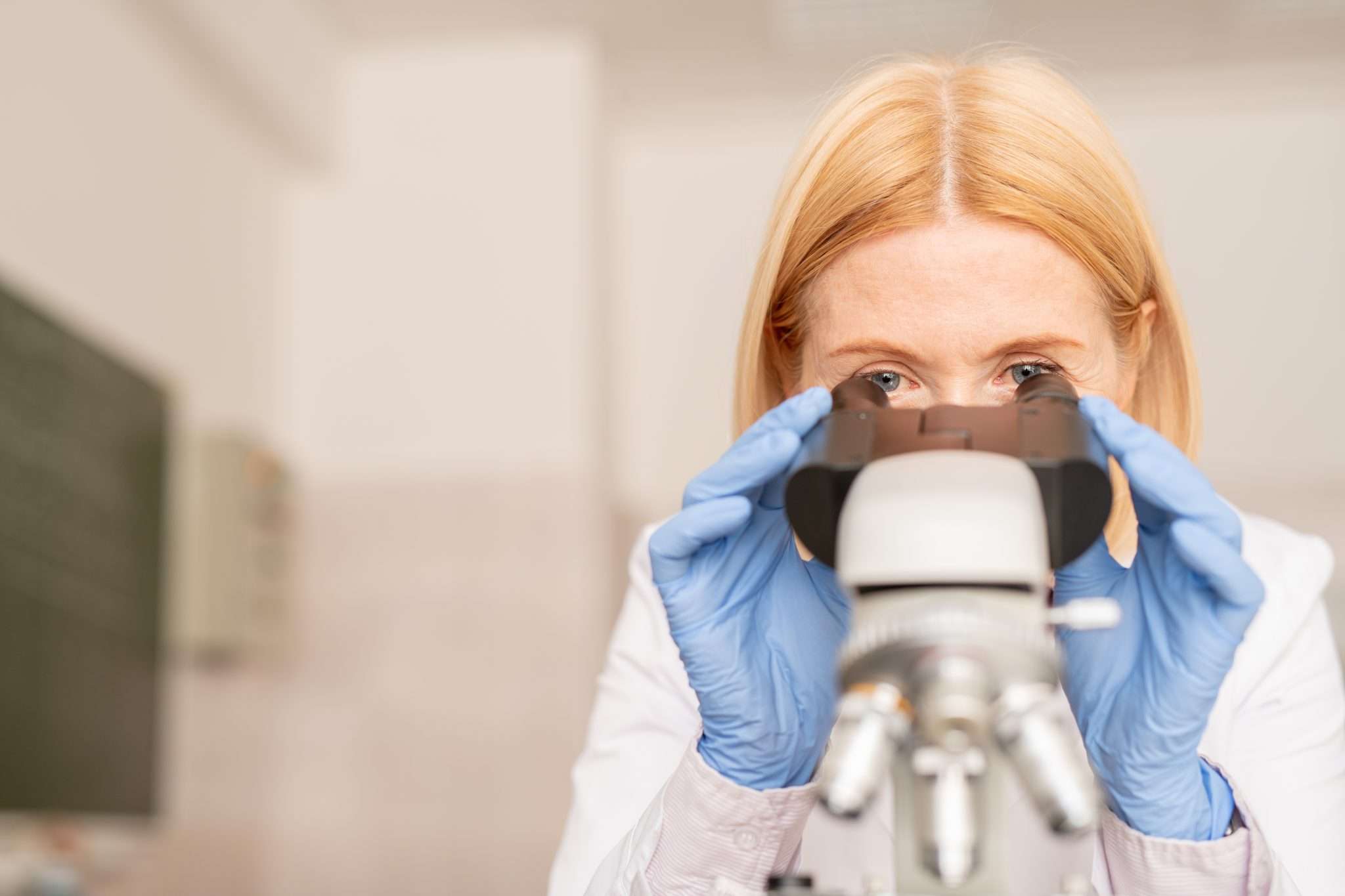 Serious concentrated blond-haired laboratory specialist in sterile gloves using microscope while doing scientific research